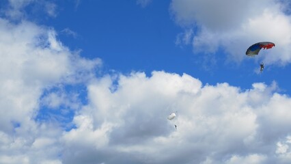 Two skydiver athletes are flying down on their parachutes against a beautiful blue sky with white...
