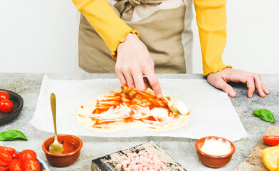 Hands of a Caucasian teenage girl smear a spoonful of pizza sauce on a heart-shaped dough.