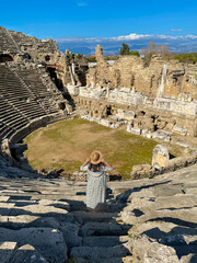 Female tourist is going down on the stairs of ancient Aspendos Theather in Antalya, Türkiye. Looking over the ruins of Aspendos with blue sky background.