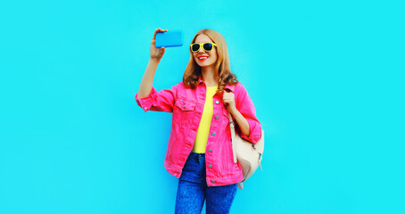 Portrait of happy smiling stylish young woman taking selfie with smartphone wearing pink jacket, backpack, yellow sunglasses on blue background