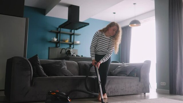 Young curly caucasian woman vacuums floor in her apartment. Adult female vacuum-cleaning the rug in modern stylish apartment. Process of cleaning the house. Housewife cleaned up the mess in the room.