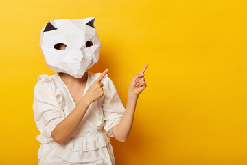 Image of unknown woman wearing dress and paper cat mask posing isolated over yellow background, pointing both fingers aside, presenting mockup for advertisement.