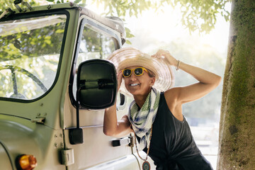 a woman wearing a hat and yellow sunglasses, looks at herself in the rearview mirror of her car. concept of well-being and happiness in middle age.