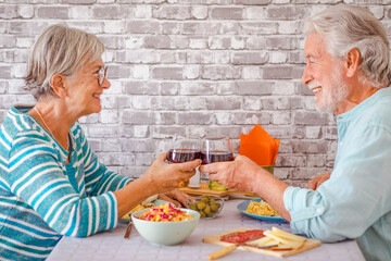 Cheerful caucasian senior couple toasting with red wineglass while sitting face to face at table having brunch or breakfast together at home
