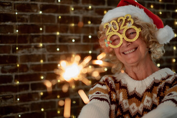 Handsome blonde senior woman with funny glasses and winter sweater holding sparkler light, elderly...
