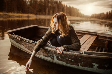 A beautiful blonde woman in a vintage sweater sits in an old wooden fishing boat floating in a...
