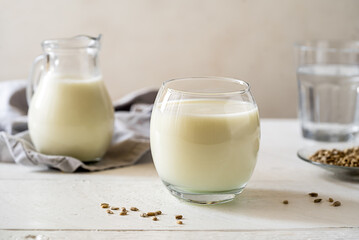 Sunflower milk in glass with raw seeds, milk jar, water, napkin on white wooden background