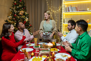 Diverse friends visiting for Christmas celebrating New Year holidays having dinner and rejoicing having fun, group of people eating and drinking wine at the table at home near the Christmas tree.