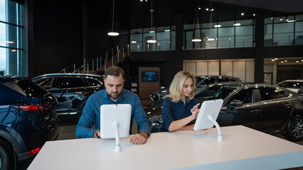 Caucasian married couple chooses a car in a car dealership on digital tablets. 