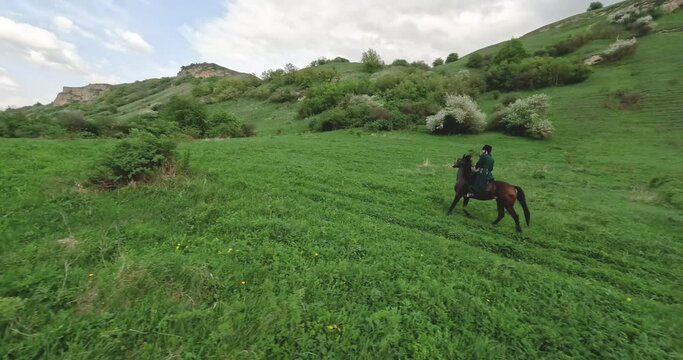 FPV sports drone shot traditional culture Caucasian horseman with gun travers riding horse at summer mountain valley landscape. Aerial view male equestrian highlander at picturesque dramatic sky rocky