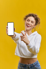 Front view of pretty girl standing with smartpone in hand. Beautiful young woman with curly hair showing screen, pointing by finger, smiling. Concept of modern technologies.