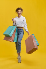 Front view of pretty girl holding packages, making step. Blonde beautiful female with curly hair posing, smiling, looking at camera. Isolated on yellow studio background.