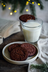 Norwegian Christmas spiced cookies pepperkaker with glass of milk and open book on wooden table and Christmas lights on background 
