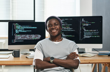 Happy young successful IT engineer in casualwear looking at you while sitting by workplace against...