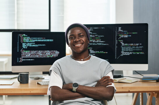 Happy young successful IT engineer in casualwear looking at you while sitting by workplace against computer screens with coded data