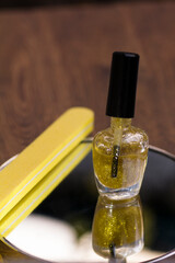 A bottle of translucent nail polish with gold glitter and a yellow nail file on a mirror surface