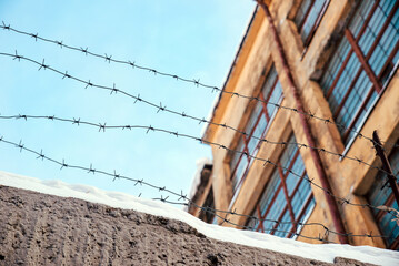 Fototapeta premium Barbed wire on a fence in snow with an old factory and blue sky on a background