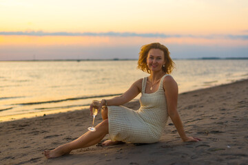 a woman with red hair flowing in the wind sits on the sand of the seashore with a glass of wine in the rays of the setting sun