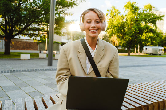 Young White Woman Using Laptop And Headphones While Sitting Outdoors