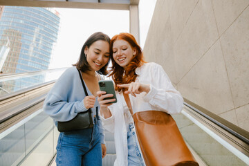 Young multinational women using cellphone while standing on escalator