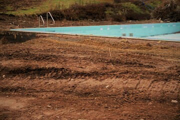Schwimmbad Panorama mit rotbrauner Erdfläche vor blauem Pool, braunen und grünen Büschen und...