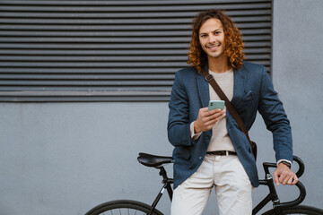 Fototapeta na wymiar Ginger man using mobile phone while walking with bicycle on city street