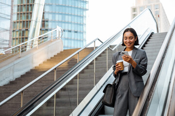 Young woman using cellphone and holding coffee while going down escalator