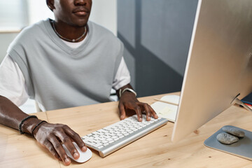 Close-up of young black man in t-shirt clicking on mouse and pressing button of computer keyboard while working in front of monitor