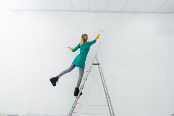 A young girl is engaged in repairs on the stairs and paints a white wall with a roller in a new apartment. Renovation of the interior and a new apartment. Housewarming and a desirable mortgage.