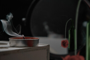A smoking candle standing on a wooden box, on the back dark background there is a round mirror in...
