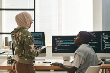 Two young intercultural IT engineers discussing ways of decoding data while Muslim woman in hijab...