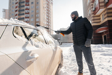 A young man cleans his car after a snowfall on a sunny, frosty day. Cleaning and clearing the car from snow on a winter day. Snowfall, and a severe snowstorm in winter.