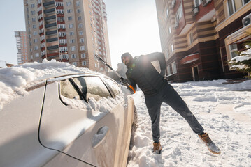 A young man cleans his car after a snowfall on a sunny, frosty day. Cleaning and clearing the car from snow on a winter day. Snowfall, and a severe snowstorm in winter. - obrazy, fototapety, plakaty