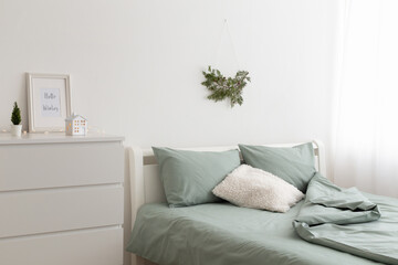 Winter bedroom decor. Christmas home decor. New year interior decorations. Coniferous branch on white wall, bed with green linen, frame with text HELLO WINTER on chest of drawers.