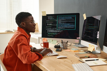 Young serious IT engineer in smart orange shirt looking at coded data on computer screens while...