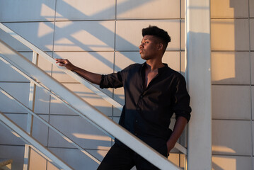 Handsome young African man wearing black shirt and pants standing on the stairs of a high building,...
