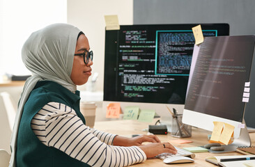 Young Muslim businesswoman in hijab looking at computer screen while typing and decoding data in...