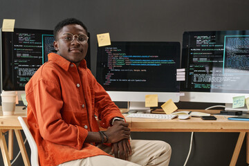 Young IT support manager looking at camera while sitting by workplace in front of computer monitors...