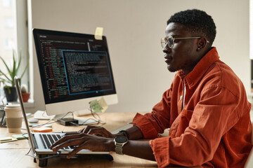 Side view of young black man in smart casualwear and eyeglasses decoding data while sitting by...