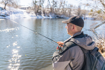 A fisherman with a fishing rod and a backpack catches fish on the bank of a snow-covered river in early spring