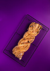 Whole handmade braided pastry with raisins, icing sugar, laminated almonds and nuts over a metallic diagonal cooling rack on a violet background.