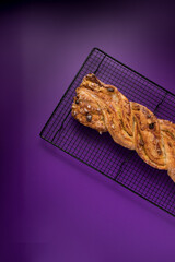 Left part of a braided handmade pastry with raisins, almonds, nuts and icing sugar over a metallic diagonal cooling rack on a purple background.