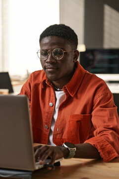 Serious young IT support engineer in casualwear and eyeglasses sitting by workplace in front of laptop and networking or decoding data