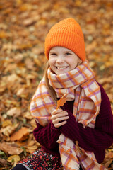 Portrait of joyful school aged girl in cute warm hat, scarf and cozy knitted sweater sitting on ground and playing with dry yellow leaves. Autumn activity, lifestyle, weekend, walking in park