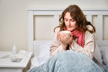 A sick woman is sitting on her home bed with a cup. Adult ill woman with a red scarf sitting on a...