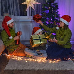 Obraz premium Happy family gives gifts at the Christmas tree in night light of the garland. Home comfort on New Year Eve in the evening living room