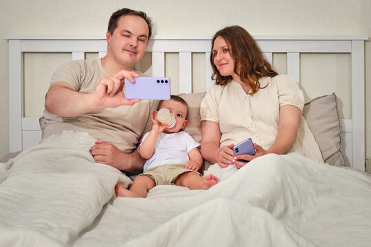 A man and a woman with a toddler baby on the bed take a selfie photo in a mobile phone. Father and mother with a child boy, happy parents and an infant baby in a home bedroom