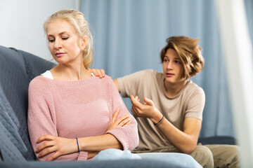 Fototapeta na wymiar Upset disappointed woman sitting at home while teenage son calming her, asking to forgive after argument ..