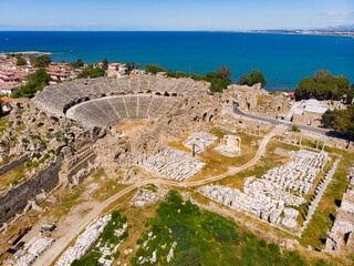 Drone view of ancient ruined Roman theater on background of Side cityscape on coast of blue Mediterranean Sea on sunny spring day. Archaeological and historical sights of modern Turkey