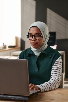 Young intercultural businesswoman in hijab and eyeglasses sitting in front of laptop and decoding data or creating new website